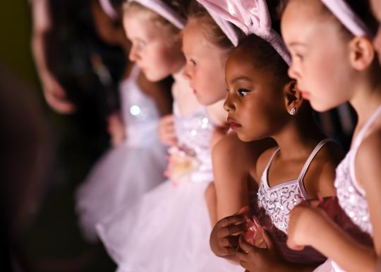 A row of ballerinas in elementary school wait on the stage for a performance.