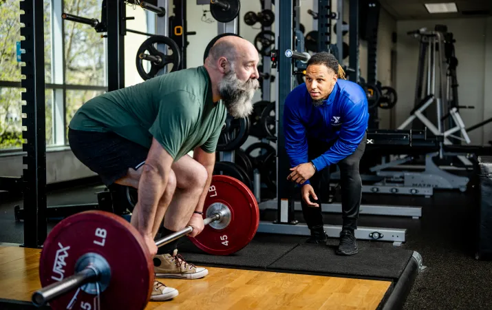 A man prepares to perform a deadlift with a Y staff member nearby in blue watching alongside for his form.