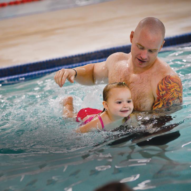A father holds his young daughter up in the water as they leans on a kickboard and creates splashes behind her. 