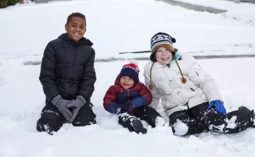 Three kids sit in the snow, two middle school age and one toddler.